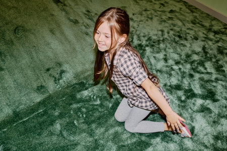 Cheerful little girl in cotton shirt and grey leggins sitting on the floor next to soft green slide after rolling down during playの写真素材
