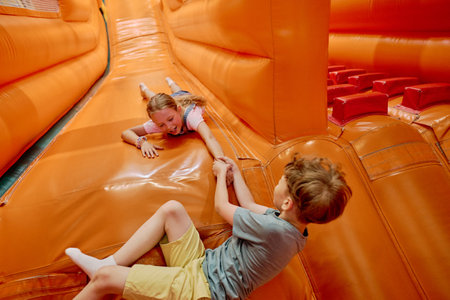Cute little pulling hand of girl while lying on top of steep orange leather slide in leisure center and having fun with friendの写真素材