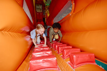 Cute little intercultural friends climbing soft orange leather tunnel with red stairs on playground...