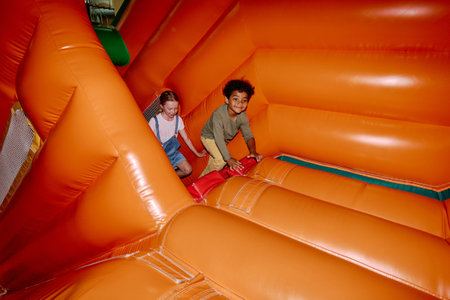 Intercultural friendly children climbing red stairs in soft orange leather tunnel while moving to...
