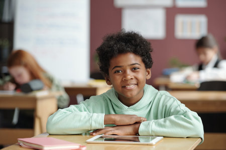 Cute schoolboy with curly hair sitting by desk in classroom with classmates on background, looking...