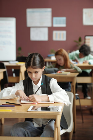 Diligent schoolgirl taking notes in copybook while sitting by desk against group of classmates at...