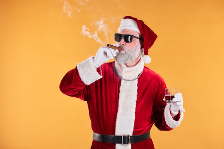 Santa Claus enjoying a cigar while holding a glass of wine in front of an orange background. Dressed in traditional red outfit, exuding cool demeanor with sunglassesの写真素材