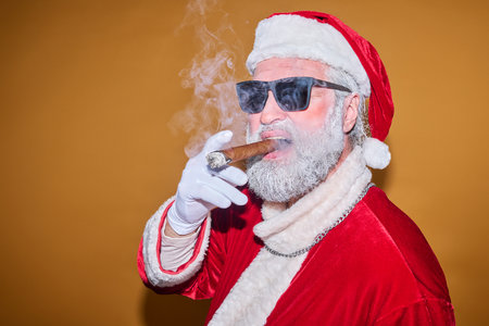 Santa Claus wearing sunglasses and smoking cigar while posing against a solid yellow-orange background with a confident expressionの写真素材