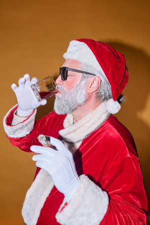 Santa Claus in traditional red suit and hat, wearing sunglasses while drinking from clear glass. Background with uniform color, creating a striking contrast with Santas attireの写真素材