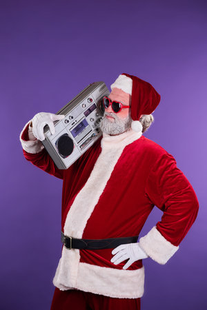 Santa Claus is dressed in traditional red suit and holding boom box on shoulder while wearing...