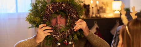 Senior man engaged in holiday tradition, holding a festive wreath adorned with greenery and ornaments, preparing for seasonal celebrations. Cozy indoor setting with warm lightingの写真素材