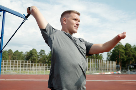 Man seen stretching with exercise band outdoors on athletic track surrounded by trees and clear sky perfect for warm-up before training sessionの写真素材