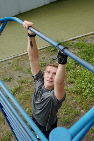 Man exercising on outdoor fitness equipment, gripping metal bars while looking determined. Athletic...