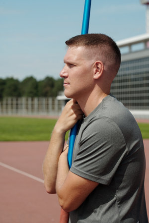 Athlete holding pole vault on outdoor track, preparing for training session. Sun illuminating...