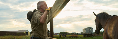 Panoramic of senior man in casual attire standing by cattle pen with brown purebred horse grazing on green grass on summer dayの写真素材