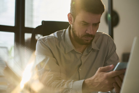 Young serious businessman looking at smartphone screen while sitting by workplace in front of laptop and textingの写真素材