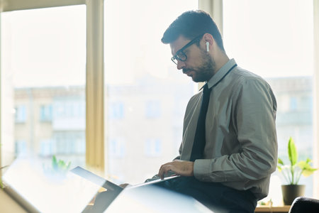 Side view of young confident market analyst sitting in front of camera in office and looking through...