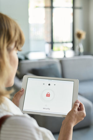 Woman holding tablet with home security application in modern living room filled with natural light...