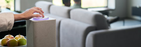 Hand adjusting settings on smart speaker against backdrop of modern living room with light furniture...