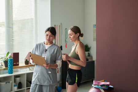 Woman in workout attire conversing with fitness professional holding clipboard at gym. Sunlight...