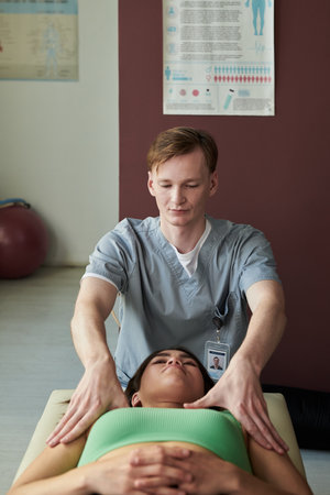 Health practitioner providing therapeutic treatment on patient lying on massage table in medical clinic setting with anatomical posters on wall and exercise ball nearbyの写真素材
