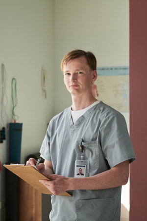 Portrait of young healthcare professional holding clipboard in medical environment while wearing...