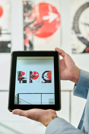 Hands of young unrecognizable male manager of art gallery holding tablet with creative artwork on screen during workの写真素材