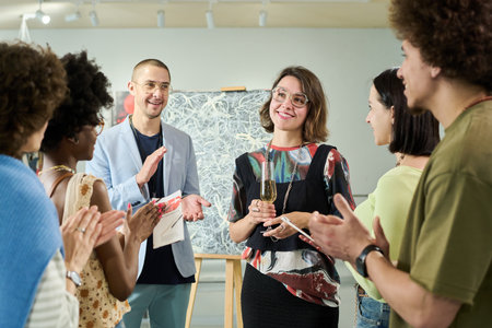 Group of visitors of art gallery applauding to happy young female artist with flute of champagne...