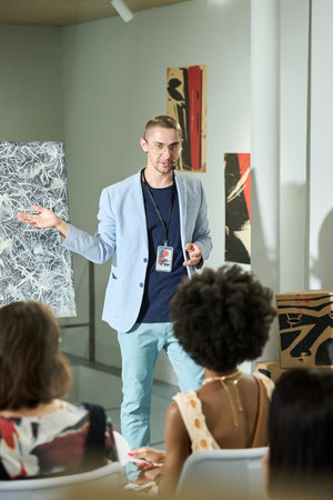 Young elegant manager of art gallery standing in front of public during presentation of new abstract...