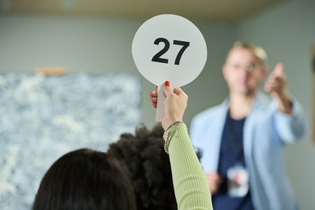 Focus on hand of young female participant of auction raising paddle with number twenty seven in front of cameraの写真素材