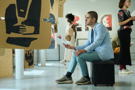Young male visitor of gallery with open brochure sitting in front of creative exposition among other...