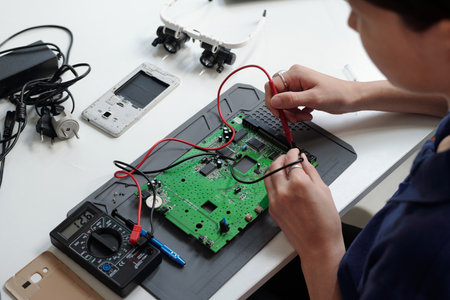 Detail of person working on electronic circuit board, using tools like multimeter and soldering iron. Various electronic components and devices are scattered on deskの写真素材