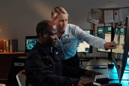 Young confident female security guard standing next to male colleague and pointing at computer...