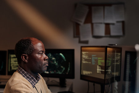 Serious mature man in beige sweater sitting in dark office and looking at computer screen while...
