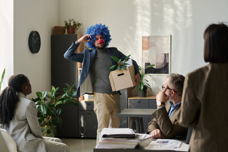 Young male clown employee with box looking at group of colleagues and keeping fingers by head while leaving officeの写真素材
