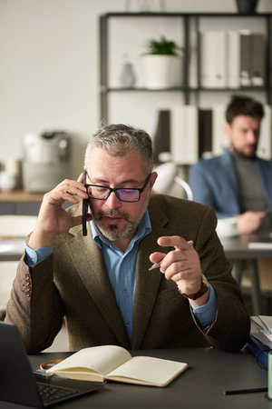 Mature businessman in eyeglasses and formalwear sitting by workplace with open notebook and speaking...