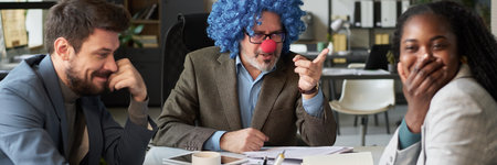 Header of young African American female employee covering mouth with hand while laughing at strict clown boss pointing at herの写真素材