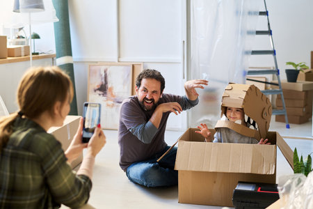 Young man imitating dinosaur while sitting on the floor next to to little daughter in box and posing for his wife with smartphoneの写真素材