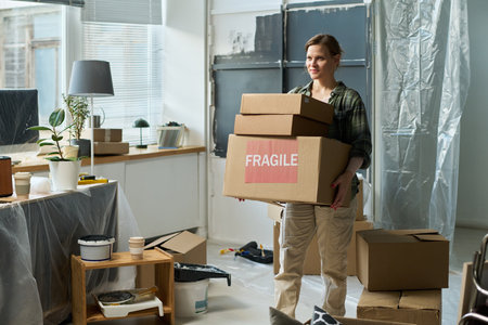 Young woman in casual attire carrying cardboard boxes with fragile household stuff while walking...