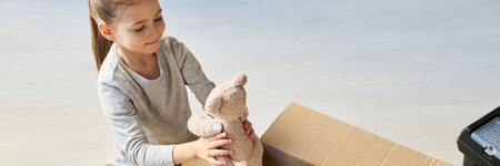 Header of little girl sitting on the floor of domestic room and unpacking cardboard box with...
