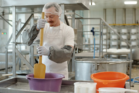 Man wearing protective gear stirring yellow mixture in large container in industrial kitchen...
