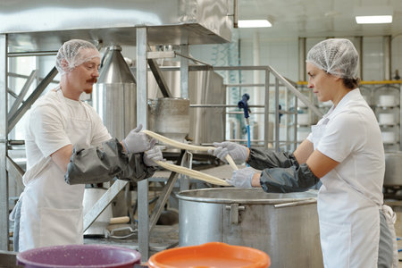 Workers preparing ingredients in large stainless steel vats in industrial kitchen wearing protective...