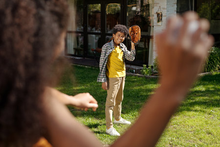 Child standing on green lawn under sunlight, holding baseball glove and smiling at camera surrounded...