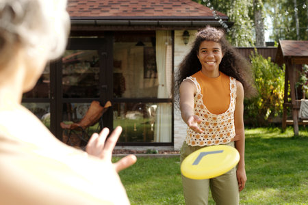 Young woman with curly hair wearing sleeveless top is playing frisbee in a backyard on a sunny day with another person whose back is turned.の写真素材