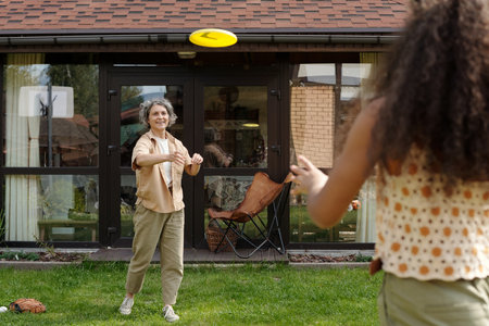 Elder woman throwing a frisbee in backyard garden, enjoying a sunny day outdoors while spending quality time with a companion, wooden furniture in backgroundの写真素材