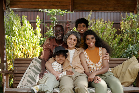 Multiracial family sitting on swing in backyard, embracing each other and smiling while spending time together outdoorsの写真素材