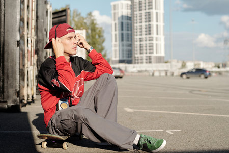 Young man in bright red hoodie adjusting his ball cap while sitting on skateboard in urban parking...
