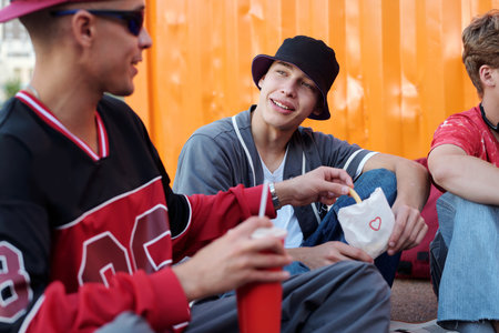 Group of young Caucasian men wearing casual outfits, sitting outdoors with snacks and drinks, chatting and laughing, creating friendly and joyous atmosphereの写真素材