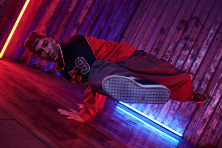 Person performing dynamic breakdance move in urban setting with neon lights illuminating wooden...