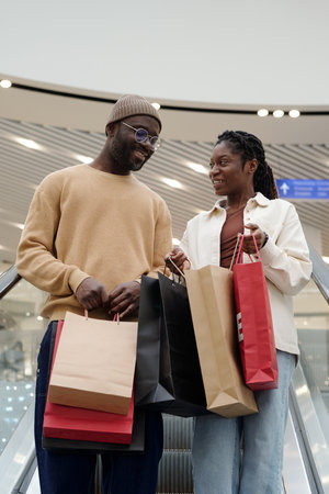 Young cheerful woman looking at her boyfriend and boasting with new purchase while opening one of...