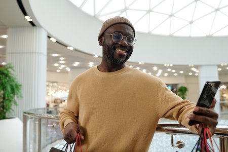 Smiling African American male shopper in pullover looking at smartphone screen after shopping while visiting trade centerの写真素材