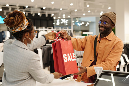 Happy young male consumer passing red sale paperbags to clerk over counter before paying for new attire in boutiqueの写真素材