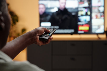 Close-up of an African American persons hand holding a remote control while watching television,...
