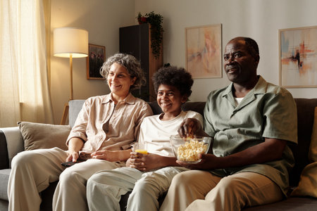Multiracial family sitting on couch and watching movie while sharing snacks in cozy living room settingの写真素材